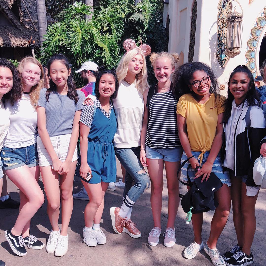 a group of eight young women and girls standing together in front of a building and foliage. they are all dressed in casual clothes, including tshirts, shorts, jeans, and dresses. one of the girls is wearing mickey mouse ears. they are all smiling and looking at the camera. the background is a mix of trees, buildings, and other people.