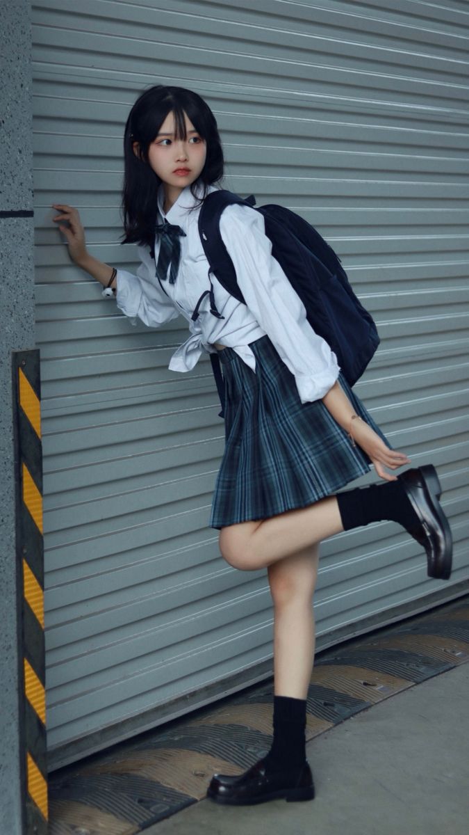 a young asian schoolgirl in a stylish school uniform stands against a gray metal wall. she's wearing a white buttondown shirt, a gray and blue pleated skirt, black socks, and black shoes.  she has a backpack on and is slightly leaning against the wall, one leg casually lifted. the overall scene is stylish and slightly edgy, with a focus on the girl's fashion and pose.