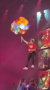 a person holding a bunch of colorful balloons in their hand with a crowd of people in front of a stage. the person is about to release the balloons.  it looks like they are at a music concert or a party.