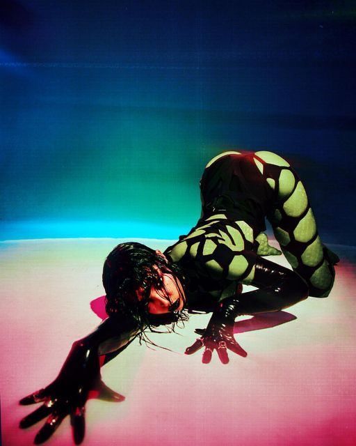 a woman in a black and green patterned latex suit is posed in a contorted position on a floor with pink and blue lighting. she is wearing several rings.