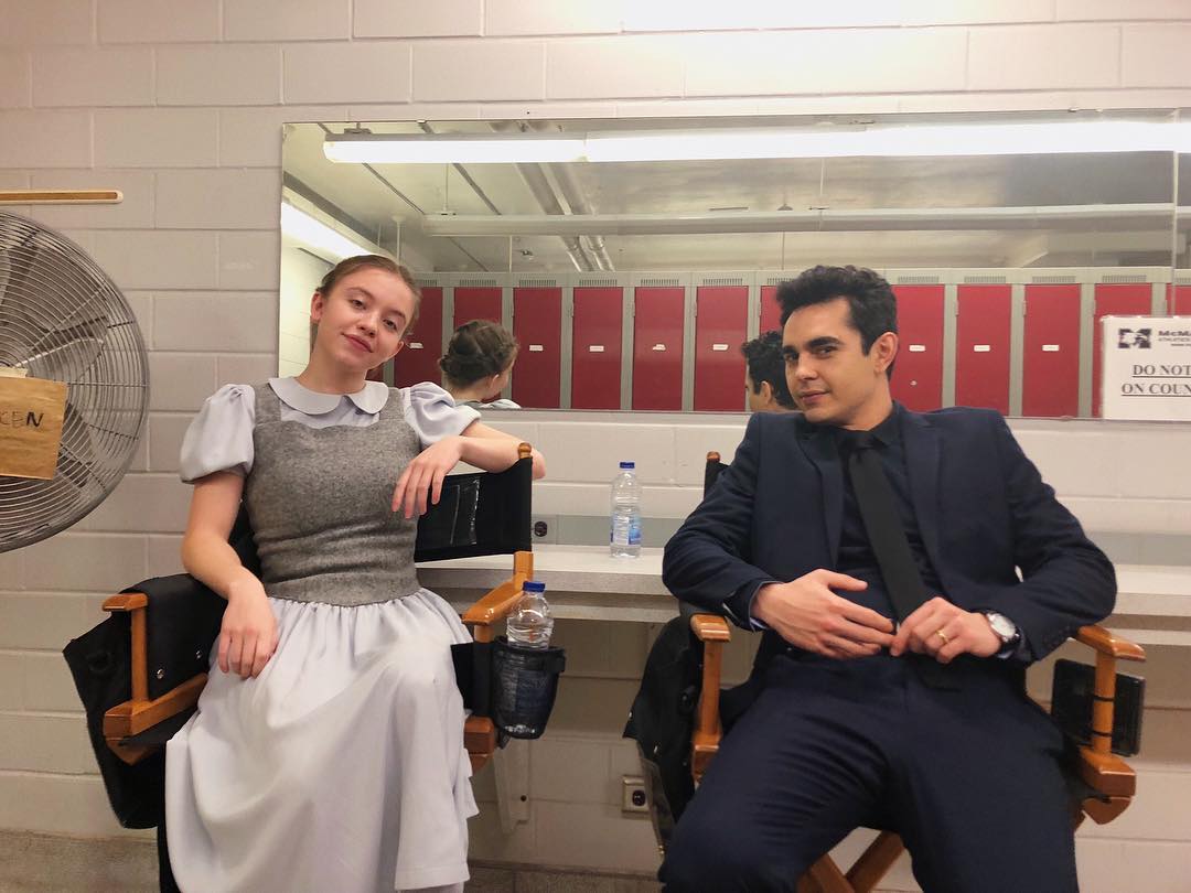 a behindthescenes shot of two young actors on a film or television set. a young woman in a light blue dress with a grey vest is seated on a director's chair, while a young man in a dark blue suit sits opposite her. they are in a locker room setting with red lockers in the background.  the overall atmosphere suggests they are preparing for a scene.