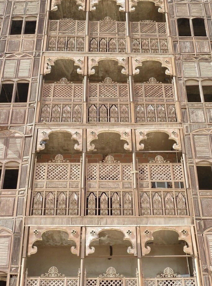a building facade with multiple levels of balconies and windows. the architectural style features intricate latticework, arched openings, and decorative details. the balconies appear to be made of stone or a similar material. the windows are a mix of closed and open shutters. the overall impression is that of traditional or cultural architecture.