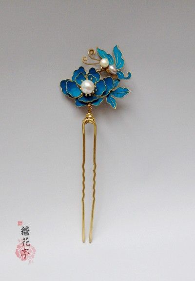 this image showcases a delicate hair stick, crafted in a traditional chinese style. the hairpin features a vibrant blue enamel flower with a dainty butterfly perched atop. the design is accented by lustrous pearls, lending an air of timeless elegance. the piece is set against a pale gray backdrop, allowing its intricate details to stand out.