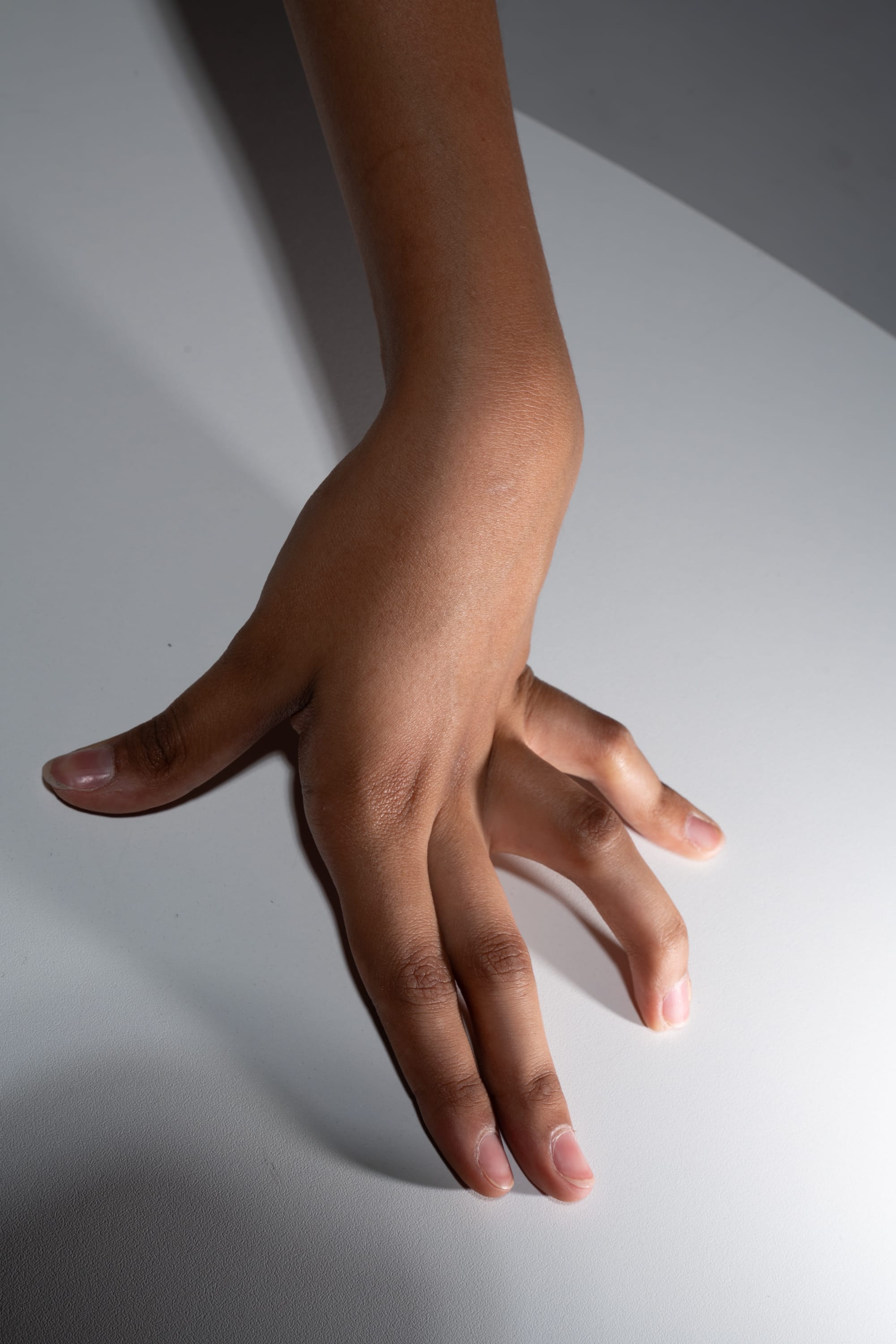 a hand with brown skin is shown with fingers spread out on a white surface. the hand is positioned with the palm facing down and the fingers pointing towards the bottom of the frame.  the background is a light gray. the image is lit with studio lighting, creating a bright and even illumination.