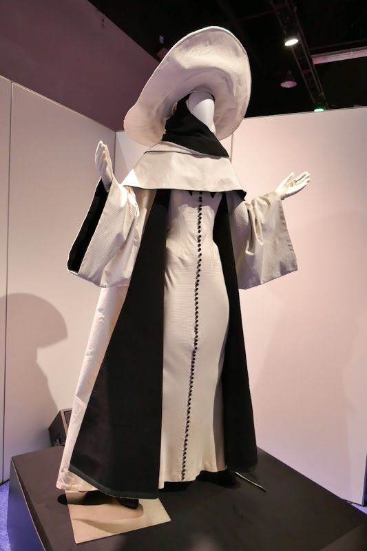 a mannequin dressed in a white and black gown with a large white hat. the dress is very formal, and the hat is large and dramatic. the mannequin is standing on a display stand in a museum or gallery. the background is plain and white, which allows the mannequin to be the focus of the image.