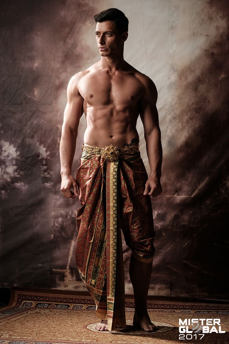 a young man stands in front of a brown background. he is wearing a traditional thai outfit, a sarong with a gold sash. he is looking to the left. the background is blurred. there is a logo on the bottom right corner that says "mister global 2017."