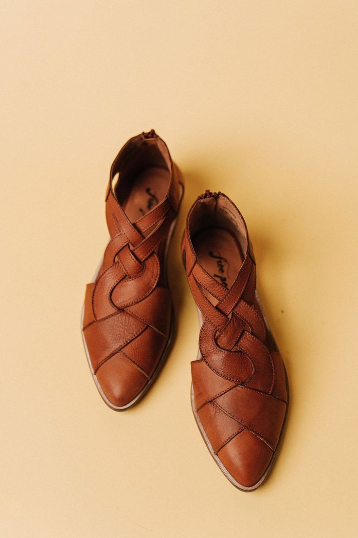 the image shows a pair of women's brown leather shoes. the shoes are a flat style with a pointed toe and feature a unique design where the leather is intricately braided and woven across the upper, creating cutouts. the shoes appear to be made of highquality leather, and the overall style is bohemian or casual chic.  they are positioned on a pale yellow or beige background, showcasing the shoes clearly. the brand name “free people” (or a similar stylized name) is faintly visible inside the shoes.