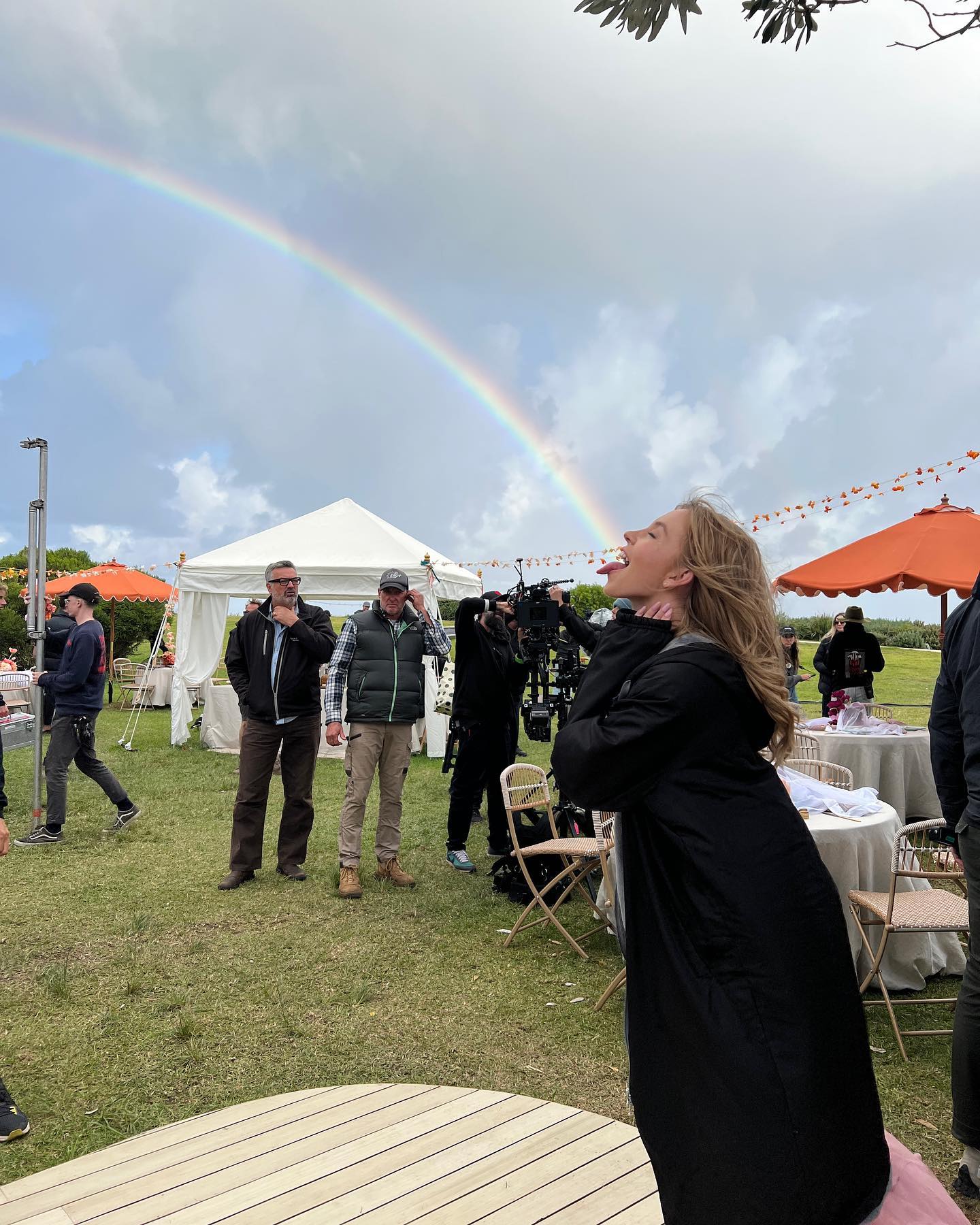 the image depicts a film set on a sunny day with a vibrant rainbow arching across the sky.  a young woman in a long black coat is positioned in the foreground, her head tilted back as if she's looking at or interacting with the rainbow. she appears to be an actress, potentially during a scene.  in the background, a film crew is visible, including individuals who seem to be the director and assistant director. they are standing around several white chairs and tables set up on the grass. there are also white tents and catering set ups suggesting a break in the filming. the overall atmosphere is one of calm and active movie production, with the rainbow adding a whimsical element.