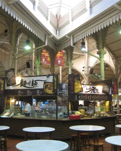 the image shows the interior of lau pa sat (telok ayer market) in singapore.  the focus is on the central food court area, with several food stalls offering various asian cuisines, predominantly chinese. the architecture is a prominent feature, with high ceilings showcasing victorianera cast iron detailing and ornate columns.  several round tables and chairs are visible, suggesting a bustling yet elegant dining environment.  the lighting is soft and creates a warm atmosphere.  a food vendor can be seen working behind one of the stalls. the overall scene conveys a vibrant and busy atmosphere typical of a traditional singaporean hawker center.