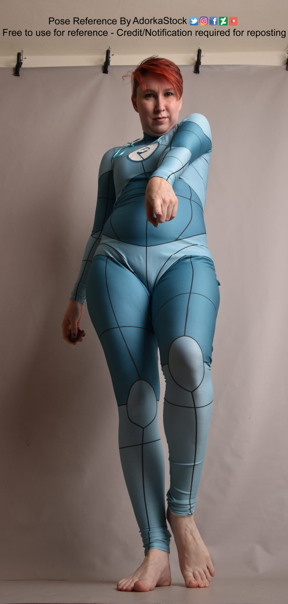 a woman is standing and posing for a reference image. she is wearing a blue and teal onepiece suit with a white trim and  is pointing her finger toward the camera. the background is plain white and the room appears to be a studio space.