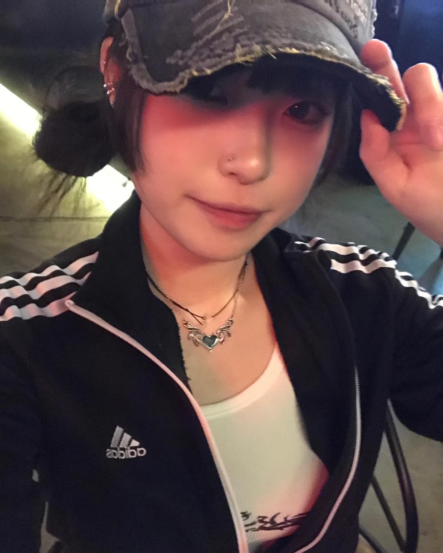 a young woman poses for a selfie indoors, likely in a restaurant. she wears a black adidas track jacket, a white graphic tee, and a black distressed baseball cap. she accessorizes with multiple earrings, a nose ring, and a silver heart pendant necklace. the lighting is primarily artificial, creating a warm and dimly lit atmosphere.