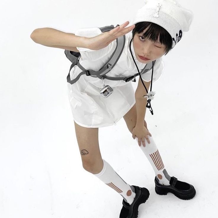 a young girl in a white dress with a black backpack, black shoes and kneehigh socks is posing for a photoshoot in a white studio. she is looking to the side.
