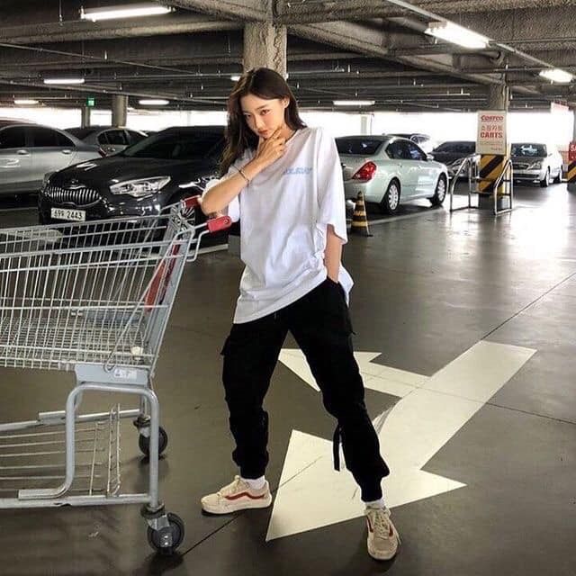 a young asian woman in casual wear poses in a parking lot. she's wearing a white tshirt, black cargo pants, and white sneakers. she is leaning against a shopping cart and looking at the camera. there are cars parked in the background. the photo is taken from a low angle and the woman's long black hair is flowing over her shoulders.