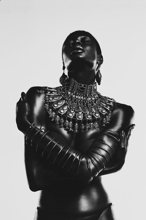 the image is a striking black and white portrait of a darkskinned female model. she is adorned with elaborate, ornate silver jewelry, including a large, statement necklace and matching bracelets that cover her arms.  the jewelry has a tribal or ethnic aesthetic. the model’s pose is powerful and slightly sensual, with her arms crossed across her chest.  the lighting is dramatic, creating high contrast and highlighting the texture of both the model's skin and the jewelry. the overall effect is one of elegance, strength, and a sense of mystery. the background is plain and white, drawing all attention to the model and her adornments.