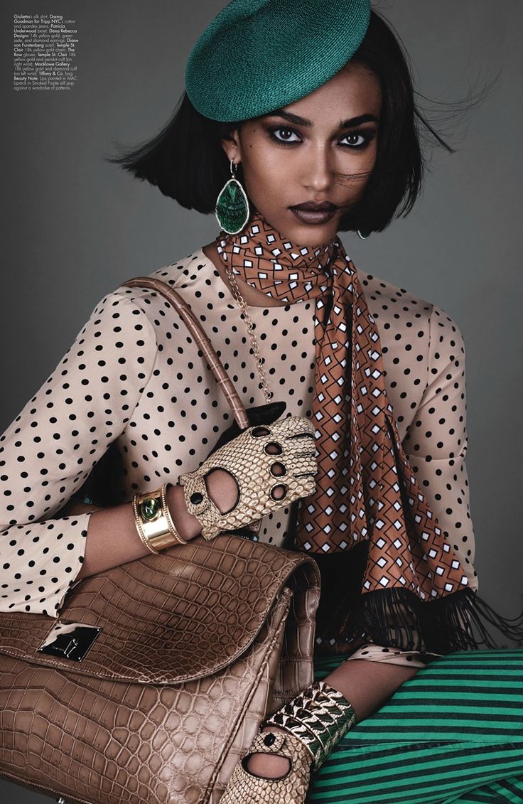 a fashion photograph of a model, showcasing a sophisticated and elegant style. she is wearing a teal beret, green earrings, a polka dot blouse, a geometric silk scarf, driving gloves, a crocodile handbag, and a gold bracelet. her makeup is dramatic, and her pose is confident. the overall aesthetic is retro and chic, suggesting a fall or winter season. the background is a simple, neutral gray.