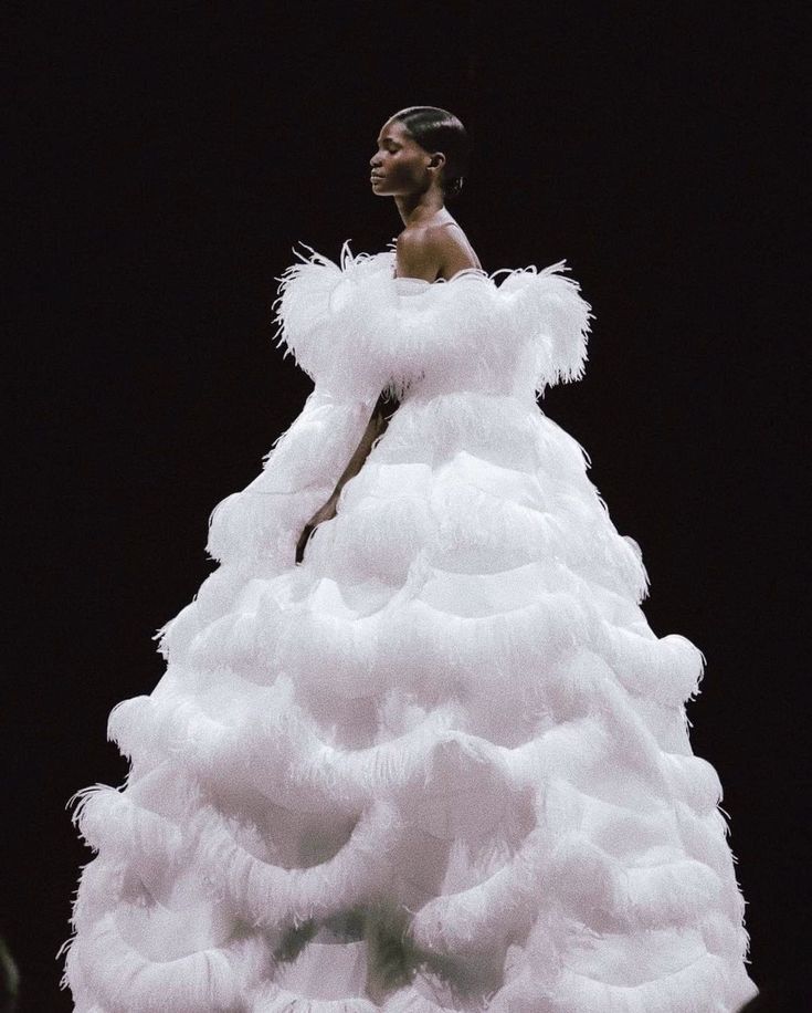 a model wearing a dramatic white feather gown on the runway. the dress has a voluminous skirt and a simple offtheshoulder bodice. she poses with her head tilted down, looking elegant and sophisticated. the dress is embellished with feathers all over, creating a textural and luxurious effect. the background is black, which creates a striking contrast with the white dress.  the overall image is one of high fashion and elegance.