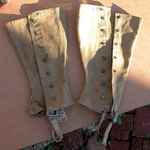two pairs of canvas military leggings are laid out on a wooden surface. they are fastened with buttons down the front and sides, and have straps that would have been used to secure them to the wearer's legs. the leggings are a tan color and are showing signs of age and use, including wear and tear and discoloration.