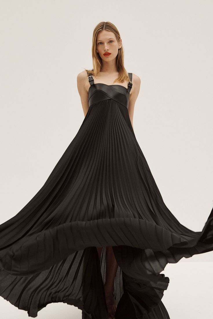 a woman is modeling a long, flowing black dress. the dress has a pleated skirt and leatherlike straps with buckles. the model is standing in a studio with a light background, and the dress is billowing around her.