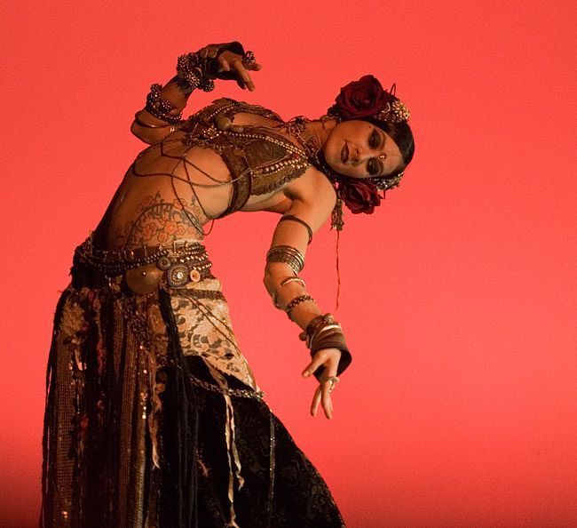 a woman in a beautiful and elaborate costume is performing a belly dancing routine on a stage. she is wearing a variety of jewelry and her expression is intense and passionate. the backdrop is a vibrant red.