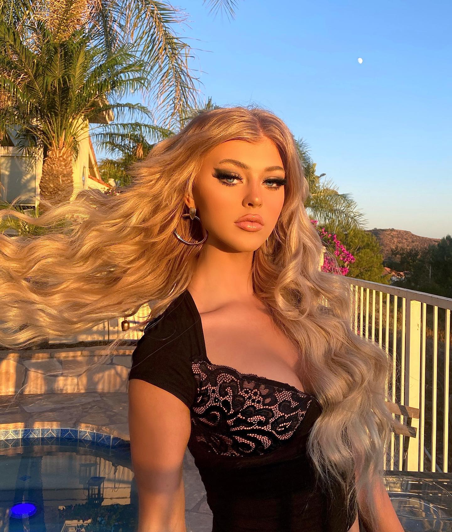 a portrait of loren gray in the evening light, standing outdoors with palm trees, a pool, and a residential area in the background. she has long blonde hair, wears a black top with lace detailing, and large hoop earrings. a full moon is visible in the sky.