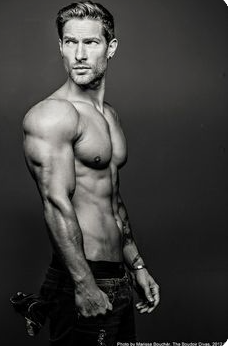 a black and white portrait of a muscular man with chiseled features standing against a dark gray background. he is shirtless, highlighting his toned physique, and his gaze is directed offcamera. he wears jeans and a bracelet on his left wrist, and his hands are partially tucked into his pockets. the image exhibits strong studio lighting that emphasizes his muscular definition.