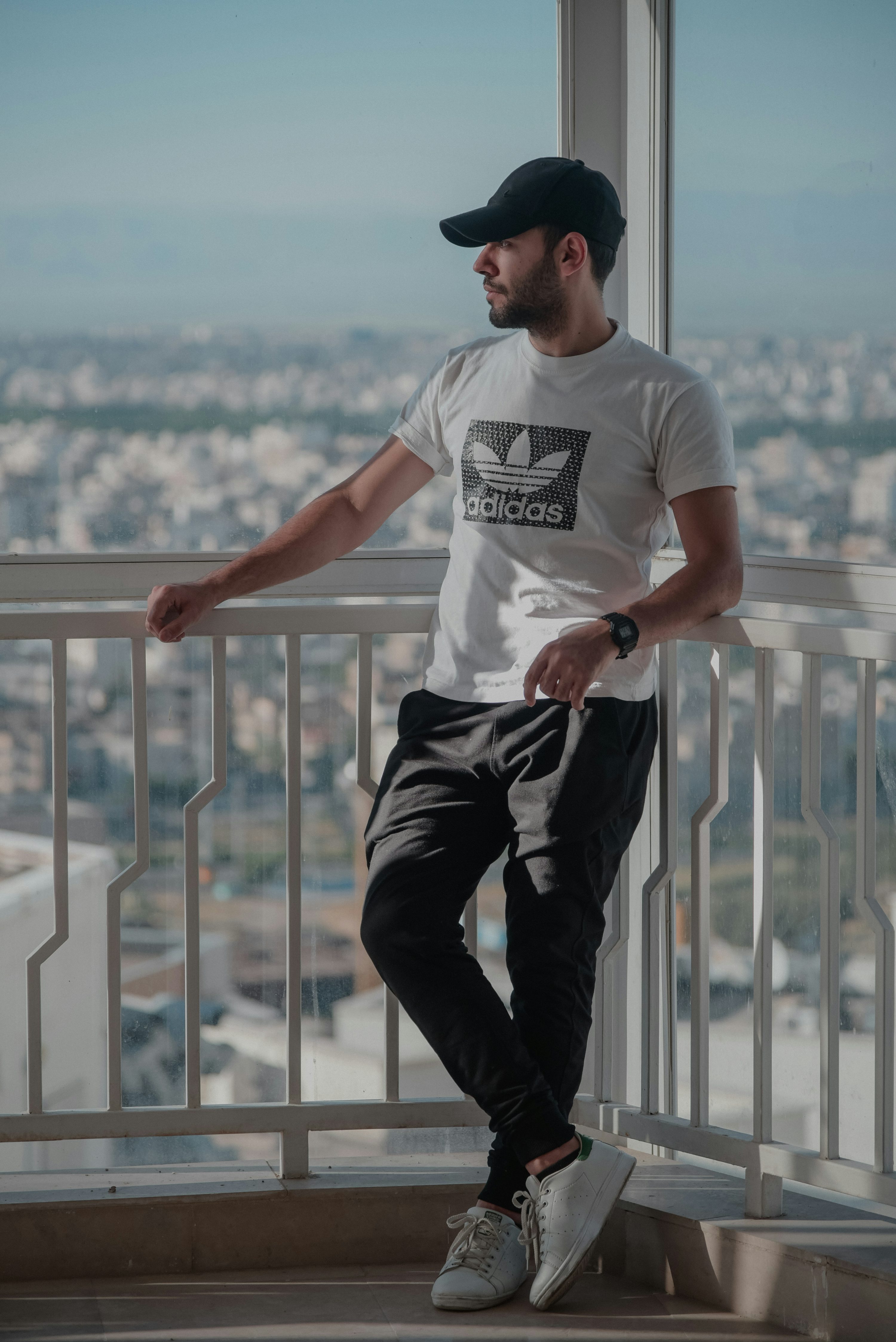 the image shows a young man standing on a balcony overlooking a city. he's wearing casual attire: a white adidas tshirt, black pants, a black baseball cap, and white sneakers. his posture is relaxed, with one leg casually crossed over the other, and he appears to be gazing pensively at the cityscape.  the setting is bright and sunny, and the overall aesthetic is clean and modern. the balcony has a white metal railing.