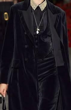 a woman in a black velvet coat and dress stands on a runway, posing for the camera. she has a black collar and a silver necklace. she is wearing a sophisticated and elegant outfit. the image captures the grandeur of a fashion show, with the focus on the woman's stylish attire. the velvet material adds a touch of luxury and the black color creates a dramatic effect. the necklace adds a subtle sparkle to the overall look. this image showcases the latest fashion trends and the allure of the runway world.