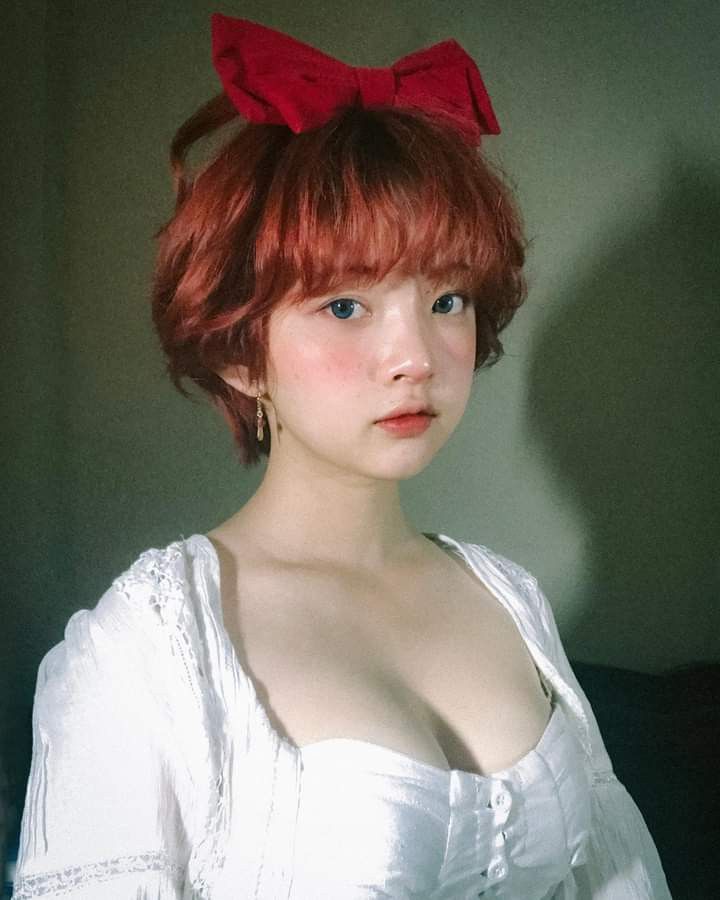the image is a closeup portrait of a young woman with short, reddishpink hair styled in a bob with bangs. she's wearing a red bow on her head and a white, offshoulder dress with a sweetheart neckline that resembles a corset top. the overall style evokes a vintage or retro feel. the lighting is soft and muted, creating a pastellike atmosphere. the woman's expression is calm and her features are delicate. the photograph has a polished and professional aesthetic, likely a studio shot designed for fashion or beauty purposes.