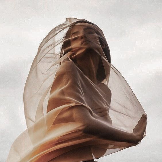 the image is a stylized portrait of a woman partially veiled by a sheer, flowing beige fabric that drapes over her body.  the fabric is translucent, allowing glimpses of her skin and form.  her expression is serene and slightly mysterious, and her dark hair blends with the fabric. the overall aesthetic is ethereal and dreamy, with soft lighting and a muted color palette creating a sense of elegance and grace. the setting appears to be outdoors, with a cloudy sky in the background. the photograph's focus is on the woman's beauty and the interplay of her body and the delicate fabric.