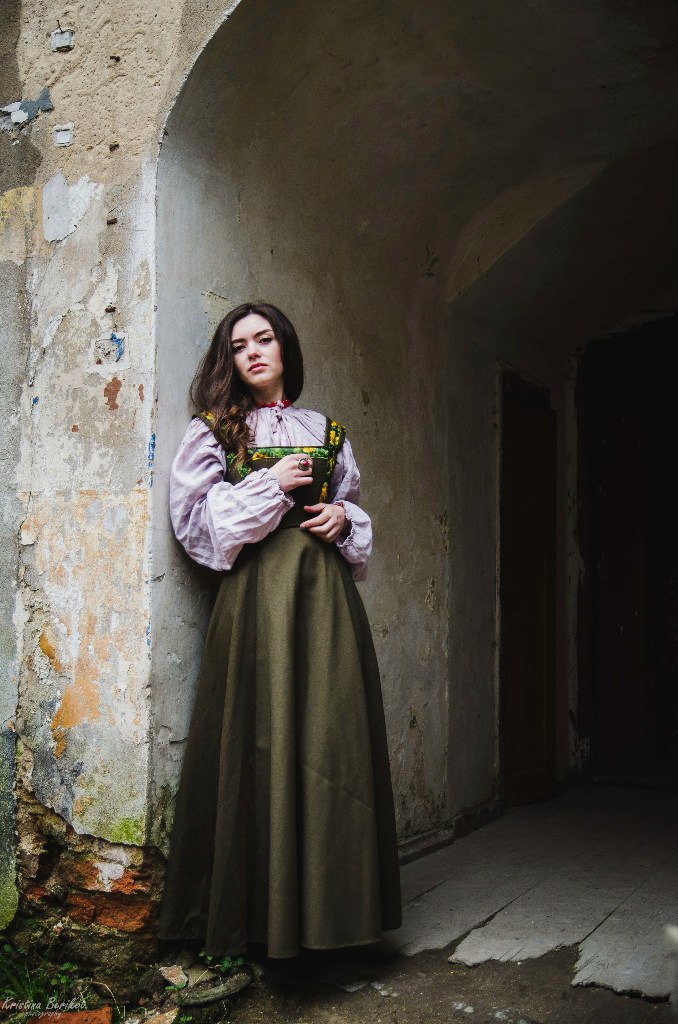 a full shot captures a woman in traditional folk attire. she is standing beside an archway, leaning slightly against a stone wall. the stone is distressed, showing signs of age. she is wearing a long, olivegreen dress, layered over a lilac blouse with long, billowy sleeves. the top of the dress features a decorated bodice with a floral pattern. her dark hair is parted to the side, cascading down her shoulder. the archway frames a dark interior, adding depth to the image.