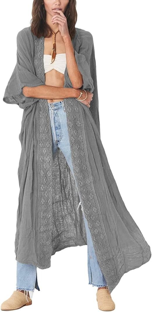 a woman is standing in a white background and wearing a gray long dress, white top and blue jeans, also wearing a brown necklace and ankle boots.