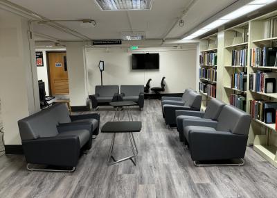 a modern, minimalist lounge area with a row of grey sofas and chairs, along with a small table, in front of a large bookshelf in the library. the room features a white ceiling with light fixtures, wood floor, large windows, and a doorway leading to another area of the building.