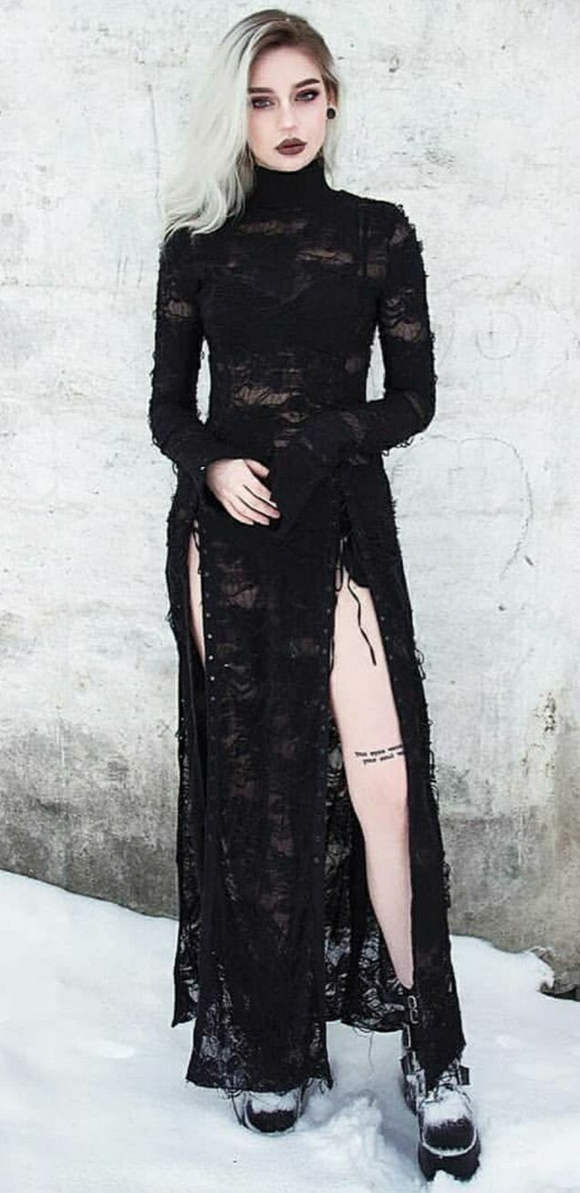 a full shot of a paleskinned woman with long, white blonde hair, wearing a black, longsleeved, lace maxi dress with side slits and platform boots. the dress is ripped and distressed, giving it a gothic aesthetic. she is posing outdoors in the snow.  her makeup is dark and dramatic. she has small tattoos on her leg.