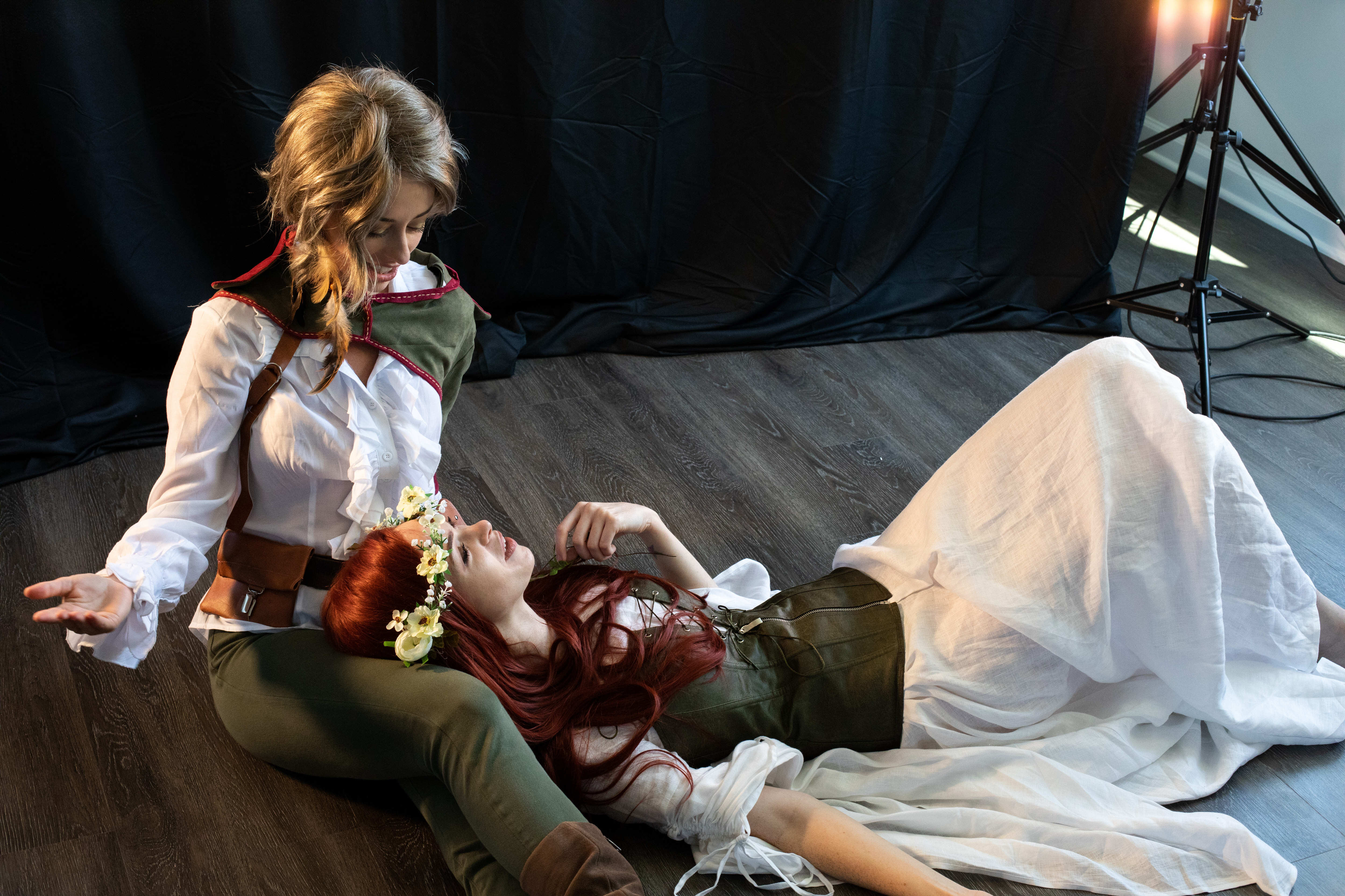 two women in fantasy cosplay are posed on a dark wooden floor against a black backdrop. one woman, dressed in white and green with a flower crown, lays across the lap of the other woman who is dressed in white and brown. the sitting woman gestures towards the camera while looking down at the other woman. they both appear happy. the lighting is soft and artificial, likely from a studio light.