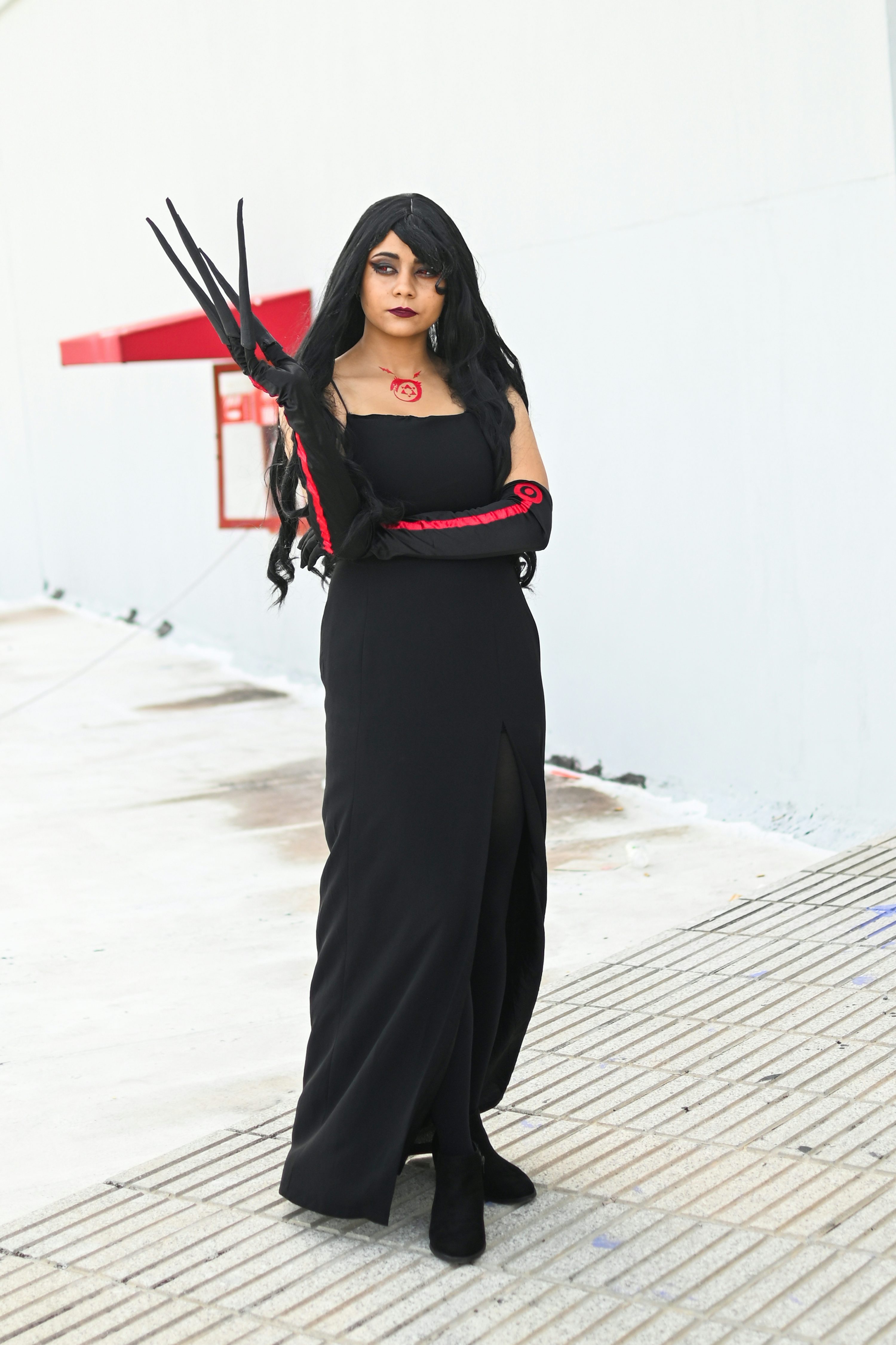 a woman is cosplaying as lucifer from soul eater. she is wearing a long black dress with red accents on the gloves and a black necklace.  she is posing with her arms crossed, her hands adorned with clawlike accessories. the background is a plain white wall and a light grey pavement. the overall aesthetic is dark and dramatic, consistent with the character.