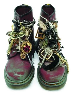 a closeup shot of two mismatched red boots heavily embellished with a variety of chains, keys, and other metal embellishments. the boots have a distressed look, giving them a vintage and punk aesthetic. the overall style is edgy and rebellious, with a focus on individual expression and creative style.