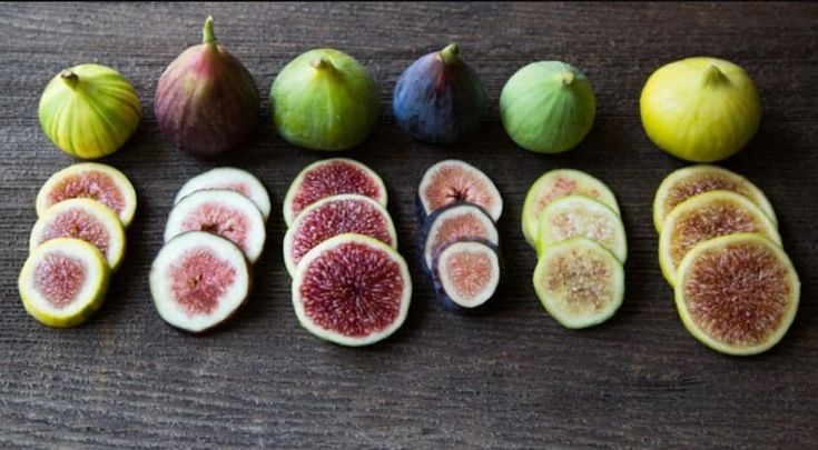 the image showcases a variety of fresh, ripe figs arranged on a rustic wooden surface. there are six whole figs of different colors (green, yellow, purple, brown) lined up at the top. below each whole fig is a small pile of slices of the same fig. the colors of the fig slices vary from light green to dark redpurple, depending on the type of fig. the photo highlights the vibrant colors and textures of the fruit.