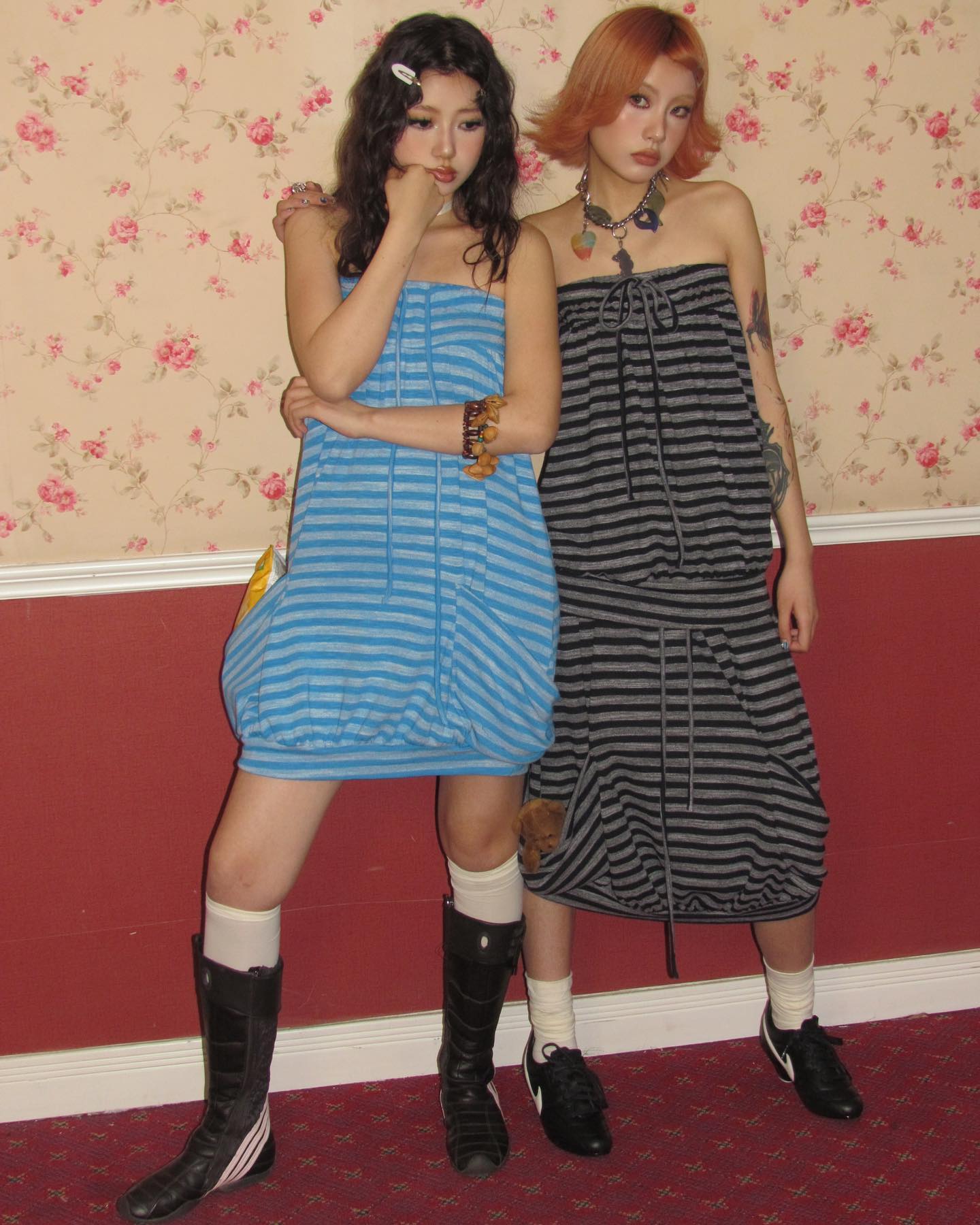 two young women are standing together against a wall with floral wallpaper.  they are both wearing striped tube dresses; one is light blue and white, and the other is dark gray and white.  they are wearing black boots and sneakers. one girl is wearing a necklace and a bracelet. they have their arms around each other and appear friendly. the background includes a red baseboard and patterned wallpaper.