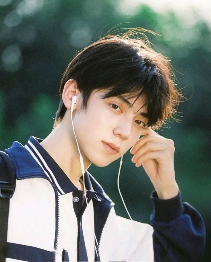the image shows a young asian man wearing a white and blue jacket. he is wearing white earphones and appears to be listening to music. he is looking directly at the camera. the background is out of focus and suggests an outdoor setting with greenery.  the lighting suggests it is taken in natural light, likely during the day.