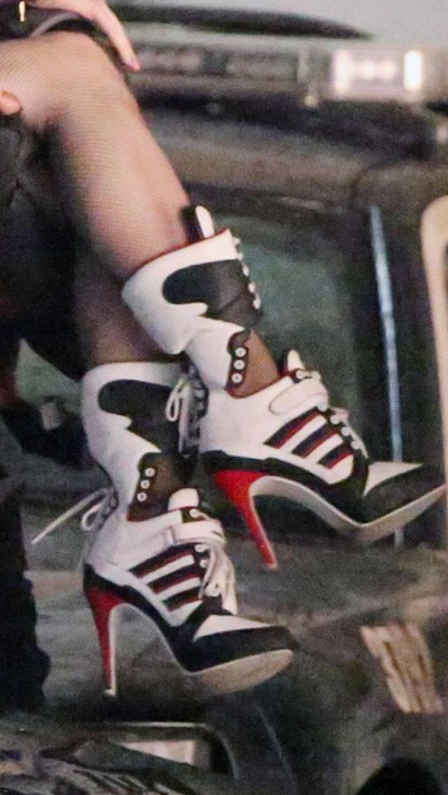 a woman in black leggings is sitting with her legs crossed. she is wearing white and black highheeled sneakers with red accents. her legs are in focus and the background is blurred. the woman is wearing a black crop top, and the image shows her lower body.  the focus is on the shoes, showing the detail and style of the high heeled sneakers.