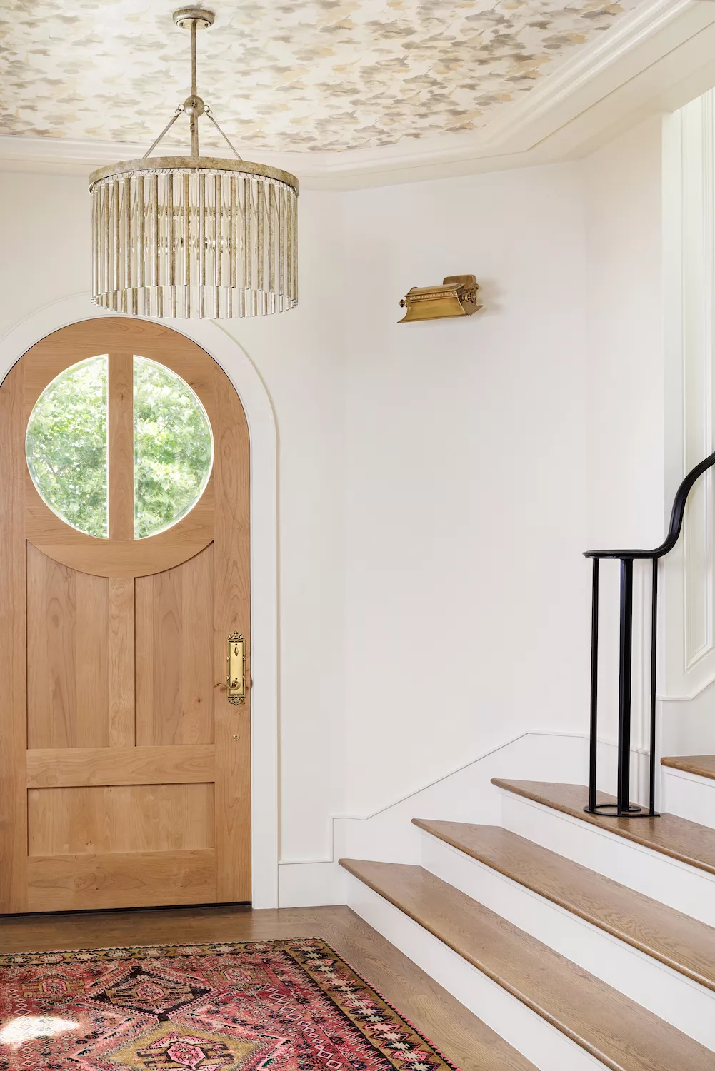 a welllit foyer showcases a staircase with a black handrail, a wooden door with a circular window, a patterned rug, and an elegant chandelier.