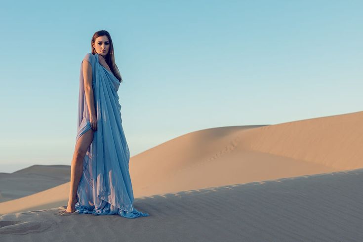 a woman in a flowing blue dress stands on a sand dune in the desert. the sun is setting behind her, casting a warm glow on the sand. she is looking at the camera with a serious expression.