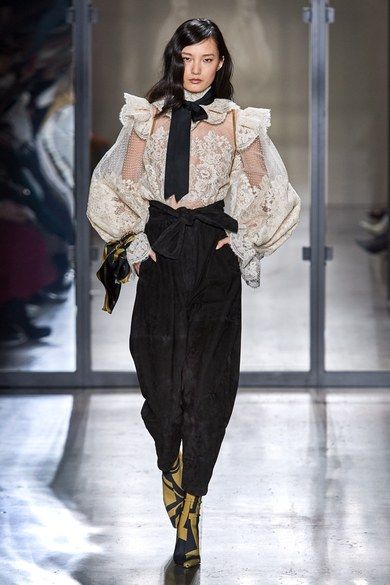 the image showcases a fashion model walking down a runway. she's wearing a striking ensemble. the centerpiece is an ivory lace blouse with voluminous sleeves, adorned with ruffles at the shoulders and a black bow tie at the neck.  underneath, she wears highwaisted, widelegged black trousers. her shoes are ankle boots with a gold and black geometric print. the overall style is elegant and sophisticated with a touch of vintage flair. the setting appears to be a minimalist runway, emphasizing the model and her attire.