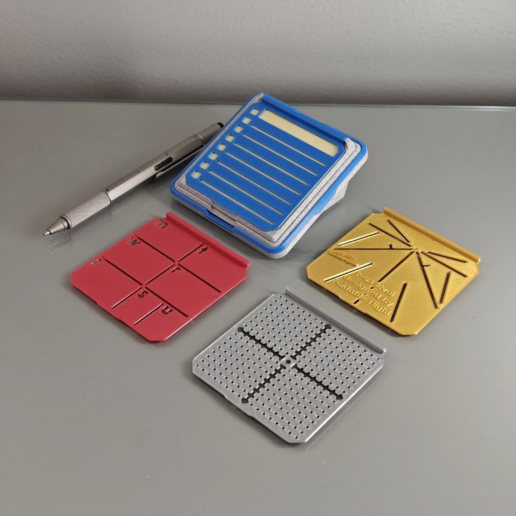 he image shows four different types of lab equipment or devices arranged on a light grey surface.  there's a pen resting near the top left.  the most prominent item is a larger, blue device with multiple slots that appears to be a sample holder or storage.  below it are three smaller, differently colored plates: a red one that seems divided into sections, a gold one with radiating lines, and a grey one with a grid pattern, likely a microplate or microarray for sample placement.  the color and design of each suggest specialized uses within scientific experiments or research. the overall impression is one of precision and careful design for scientific applications.
