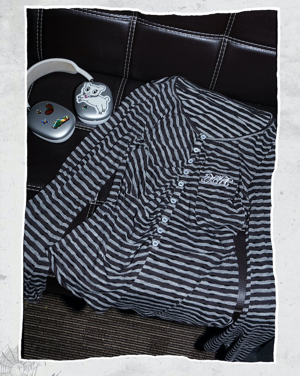 a closeup shot of a grey and black horizontally striped longsleeve henley shirt casually draped over a dark brown leather couch.  a pair of grey headphones with cartoonish stickers sits on the couch next to the shirt. the overall aesthetic is relaxed and casual.