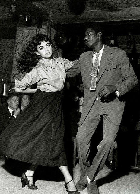 a black and white photo of a man and woman dancing in a nightclub. the man is dressed in a suit and tie, while the woman is wearing a dress. they are both smiling and appear to be enjoying themselves. the photo has a vintage feel and appears to have been taken in the 1950s. the man is african american and has a dapper look. the woman is caucasian and has long hair.