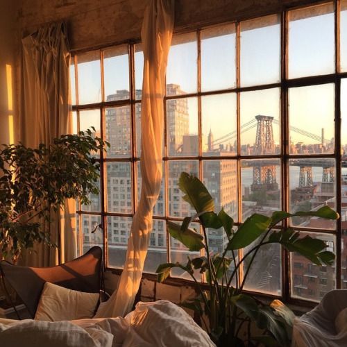 the image showcases a cozy loft apartment bathed in the warm, golden light of a setting sun.  a massive window dominates one wall, offering a stunning view of the new york city skyline, including a portion of the manhattan bridge.  the room is tastefully decorated with lush indoor plants, sheer curtains that softly filter the sunlight, a comfortable armchair with pillows, and a bed with bedding. the overall ambiance is serene, peaceful, and inviting, with a distinct aesthetic that blends urban chic with a touch of nature.  the golden hour lighting enhances the warm, inviting feel of the space.