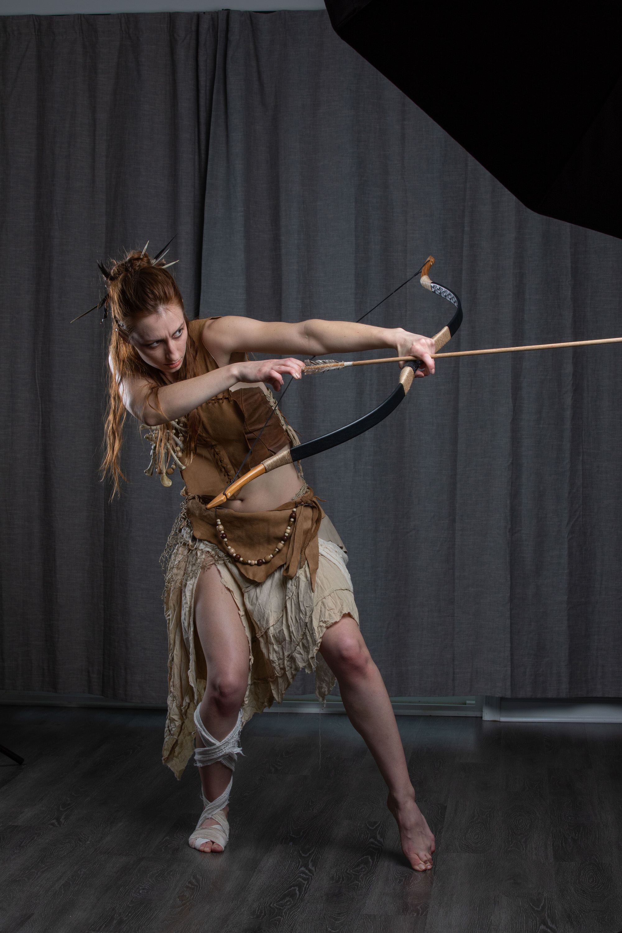 a woman with reddish hair pulled back in a bun stands in a dynamic pose, aiming a recurve bow and arrow. she is dressed in a costume suggestive of a warrior or huntress, with a leather and cloth top and skirt, and bandages on her leg. the background is a dark gray studio backdrop, and she is lit with studio lighting.