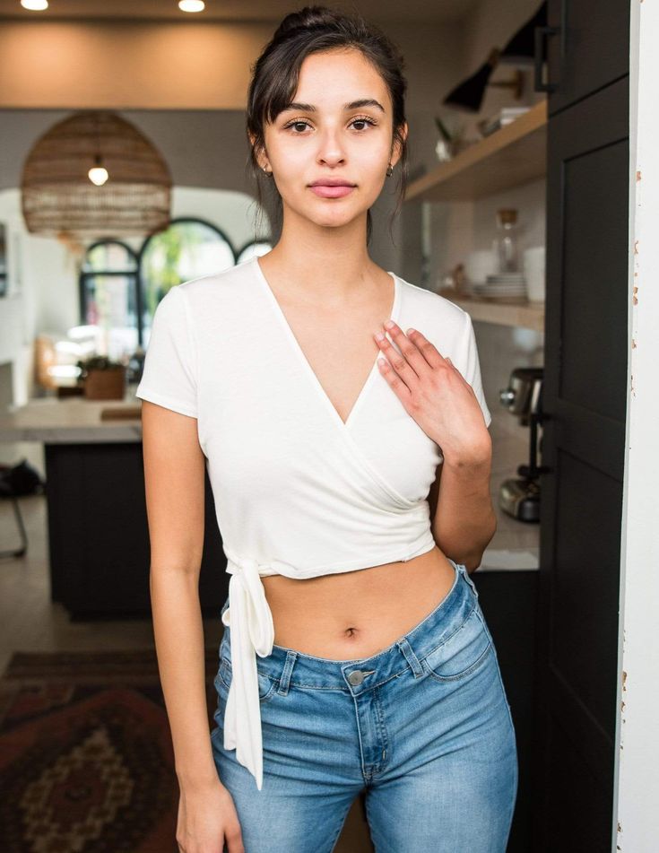 a young woman with olive skin and dark hair pulled back stands in a kitchen. she is wearing a white wrap top and blue jeans. she is posing with one hand lightly on her chest and looking directly at the camera. the kitchen is modern with white countertops and wooden accents. the lighting is bright and natural, coming from a window in the background.