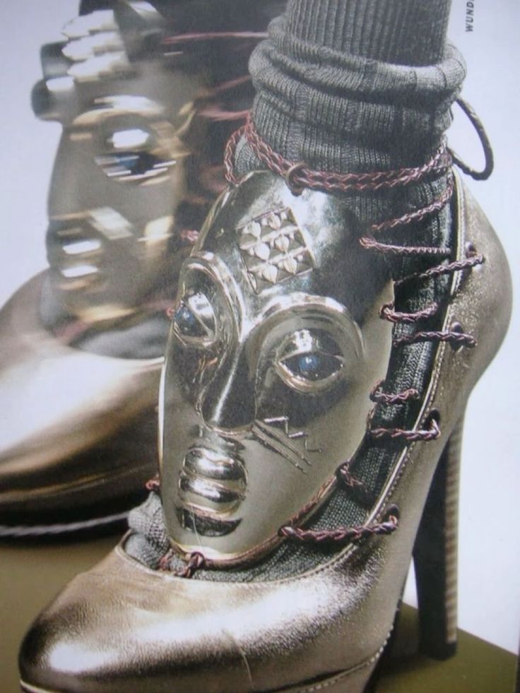 a closeup shot of a woman's foot wearing a pair of silver highheeled shoes, showcasing the unique detail of a metal mask embellishment attached to the shoe. the shoe's intricate design is emphasized by the rich texture and metallic sheen. the focus is on the shoe itself, highlighting its craftsmanship and artistic appeal. the image evokes a sense of sophistication and style, suggesting a highfashion editorial or runway context.